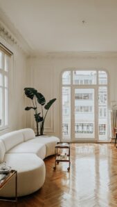 Interior of spacious light flat with couch near table and potted green plant next to windows with glass door leading to balcony